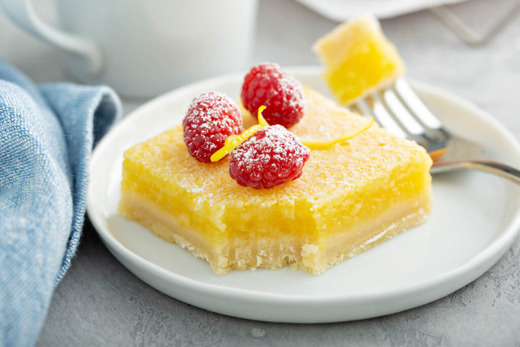 Eine angebissene Zitronenschnitte auf einem weißen Teller, im Querschnitt sind der helle Mürbeteig-Boden unten und die gelbe Zitronencreme oben zu sehen, garniert mit Puderzucker, drei frischen Himbeeren und einer feinen Zitronenzesten-Spirale, daneben eine Gabel mit einem Stück