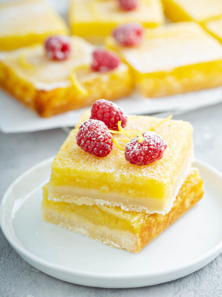 Zwei übereinandergestapelte Zitronenschnitten auf einem weißen Teller mit deutlich sichtbaren Schichten aus hellem Mürbeteig-Boden und gelber Zitronencreme, mit Puderzucker bestäubt und mit frischen Himbeeren und feinen Zitronenzesten-Spiralen garniert