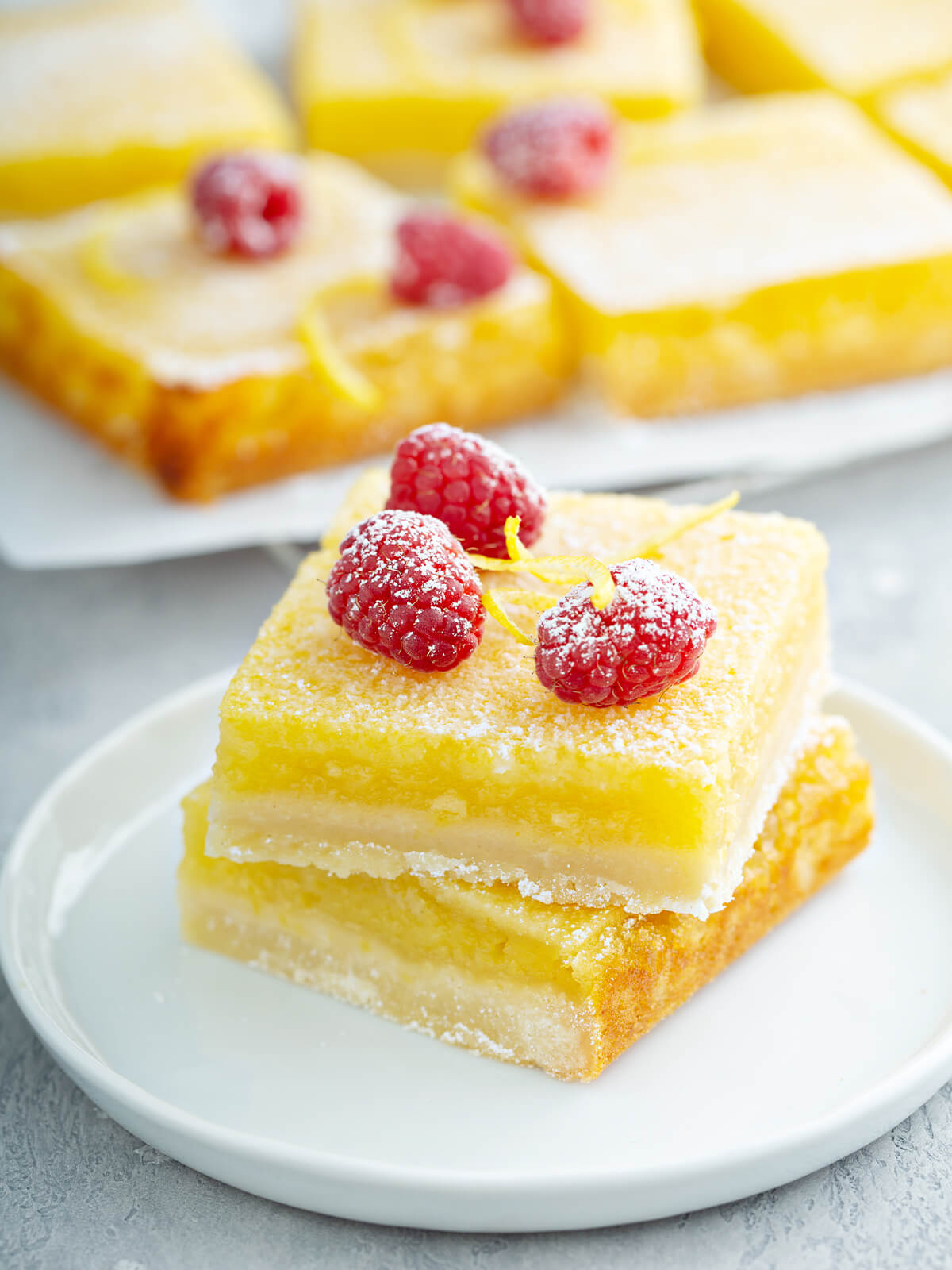Zwei übereinandergestapelte Zitronenschnitten auf einem weißen Teller mit deutlich sichtbaren Schichten aus hellem Mürbeteig-Boden und gelber Zitronencreme, mit Puderzucker bestäubt und mit frischen Himbeeren und feinen Zitronenzesten-Spiralen garniert