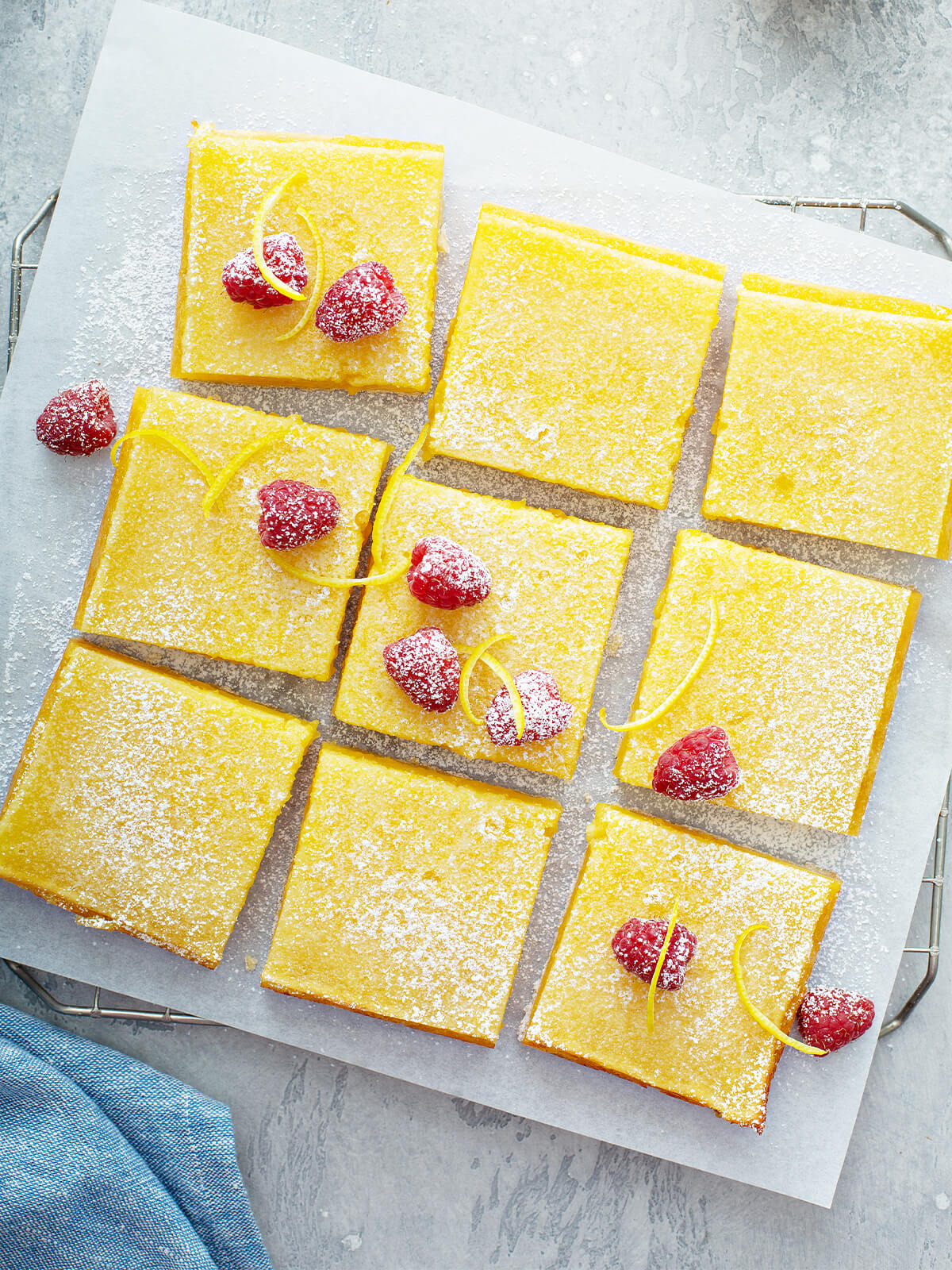Aufsicht auf neun frisch geschnittene Zitronenschnitten auf Backpapier auf einem Kuchengitter, mit Puderzucker bestäubt und dekoriert mit frischen Himbeeren und spiralförmig geschnittenen Zitronenzesten