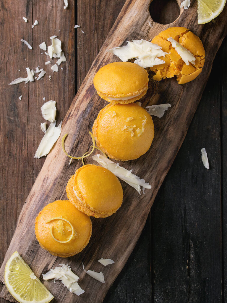 Fertige Zitrus-Macarons in einer Reihe auf einem Holzbrett, mit weißen Schokoladenspänen und Zitronenschnitzen