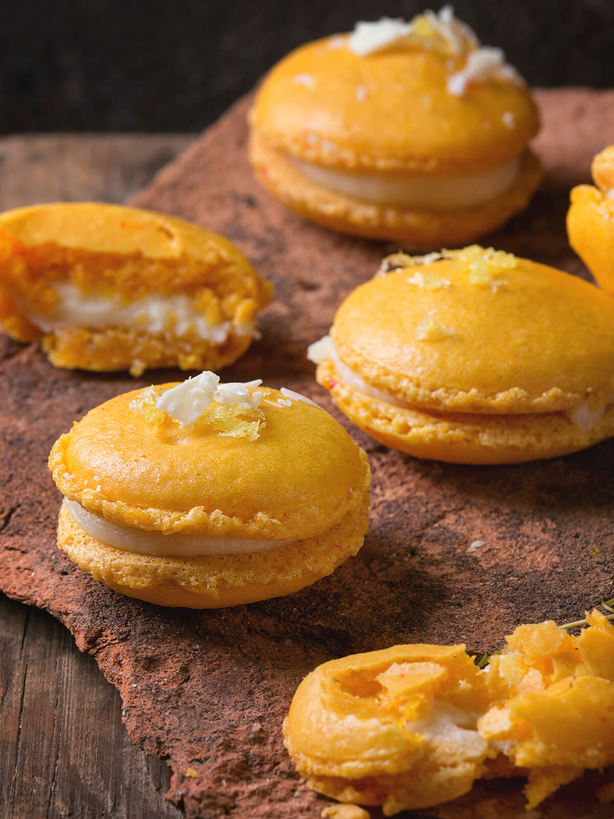 Zitrus-Macarons mit weißer Schokoladenfüllung auf rustikaler Tonplatte, garniert mit kandierter Zitronenschale