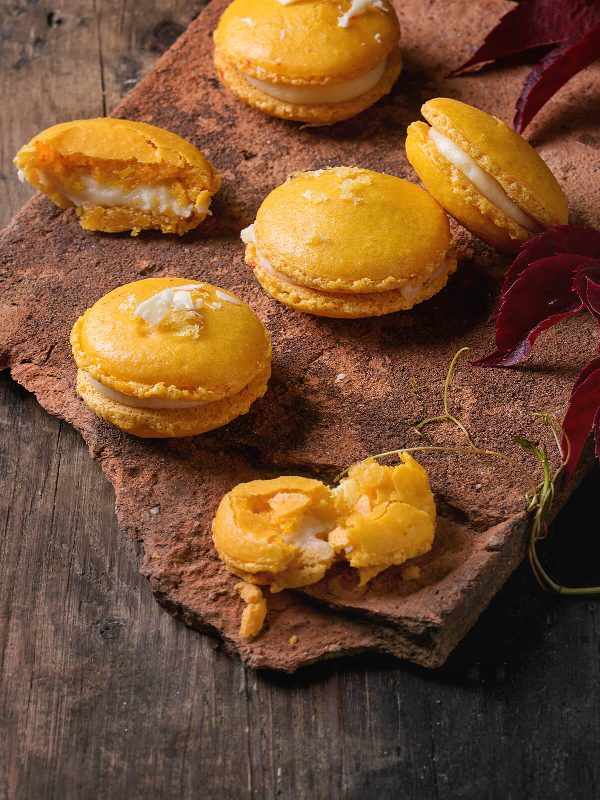 Zitrus-Macarons auf Tonplatte mit herbstlich roten Blättern, einige aufgebrochen mit sichtbarer weißer Füllung