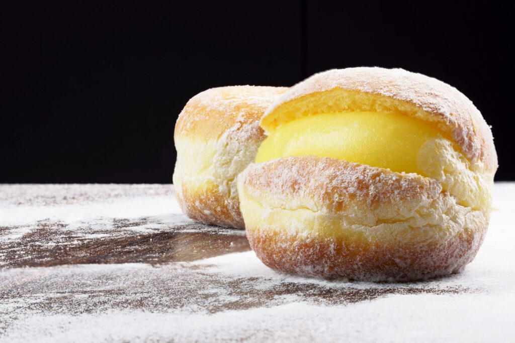 Zwei fertige brasilianische Cremekrapfen mit Puderzucker auf dunklem Holz, einer mit sichtbarer Vanillecreme