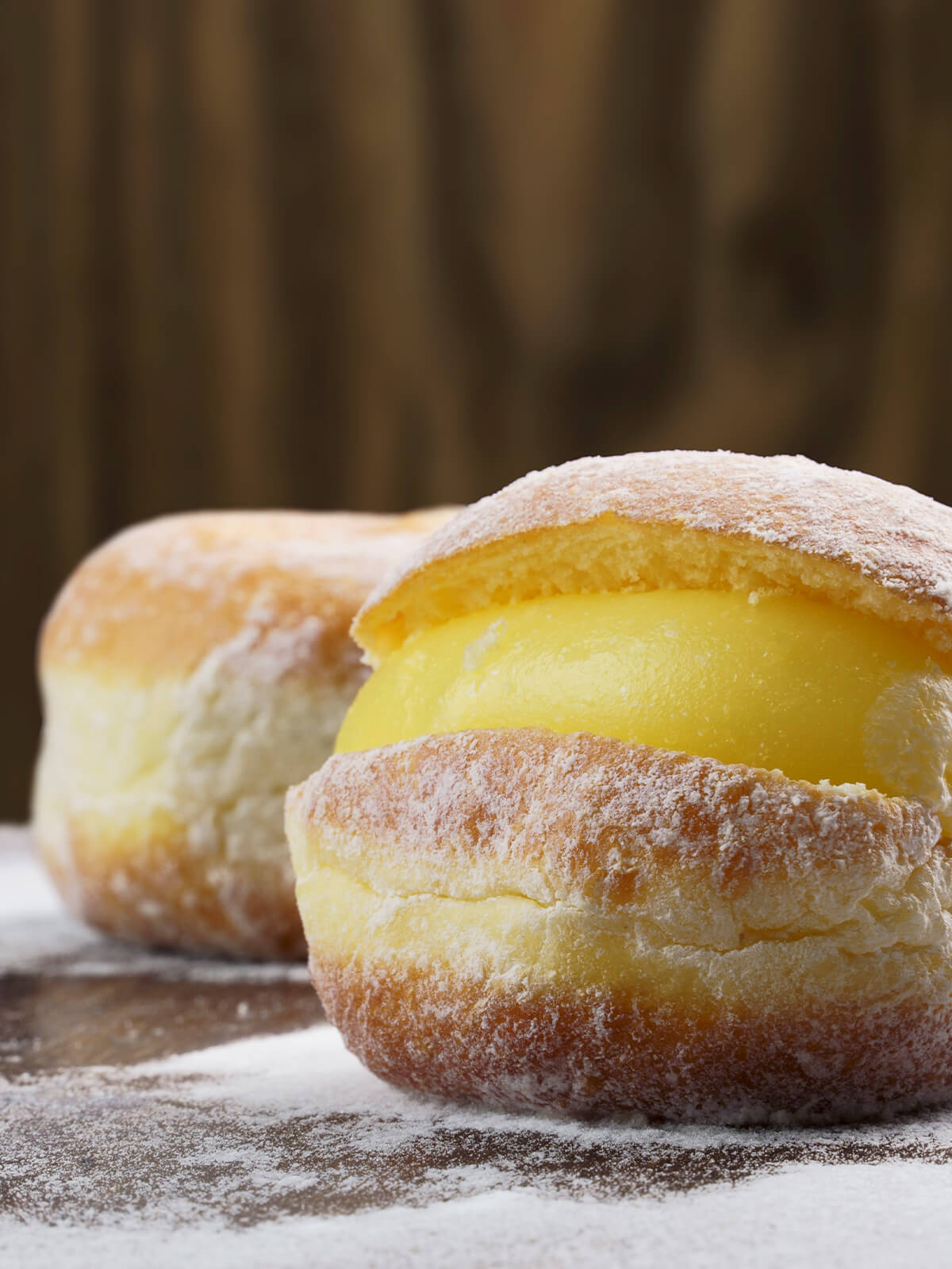 Zwei brasilianische Cremekrapfen mit Puderzucker, der vordere aufgeschnitten und mit gelber Vanillecreme gefüllt