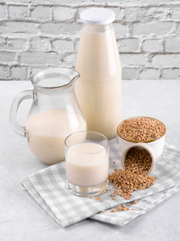 Flasche, Krug und Glas mit heller Buchweizenmilch neben zwei Schalen mit grünem Buchweizen
