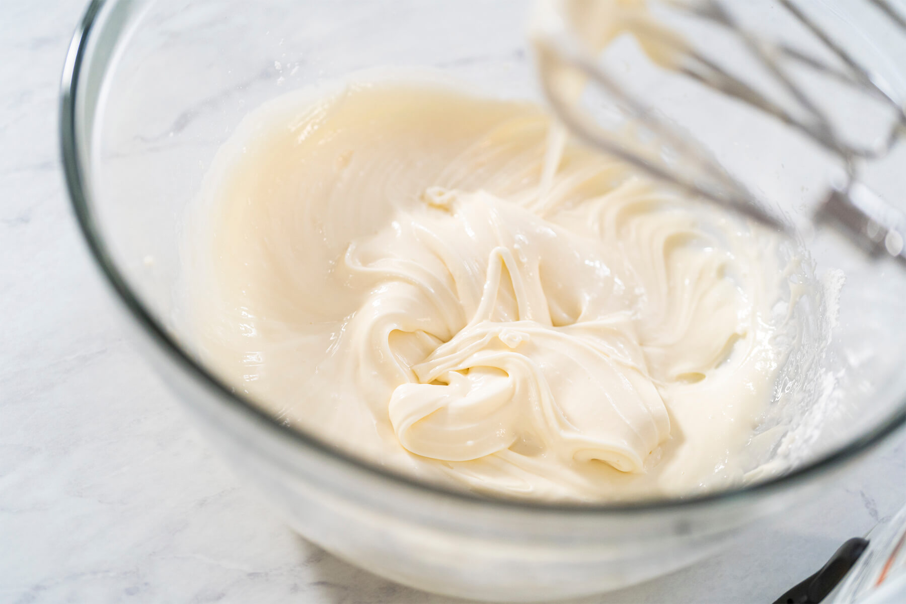 Glatt gerührte Frischkäse-Glasur in einer Glasschüssel mit den Quirlen des Handrührgeräts