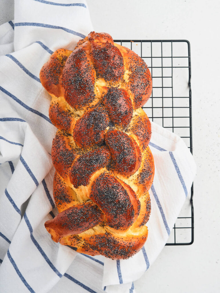 Geflochtener Hefezopf mit Sauerteig-Anstellgut, dicht mit blauem Mohn bestreut, auf einem Kuchengitter mit gestreiftem Leinentuch