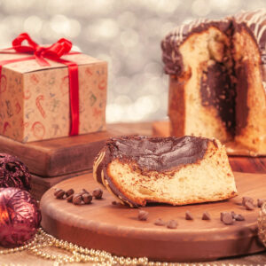Scheibe eines gefüllten Panettone mit dickem dunklem Schokoladenkern auf einem Holzbrett, dahinter der angeschnittene Laib mit glänzender Glasur