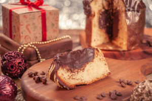 Scheibe eines gefüllten Panettone mit dunklem Schokoladenkern auf einem Holzbrett, dahinter der angeschnittene Laib