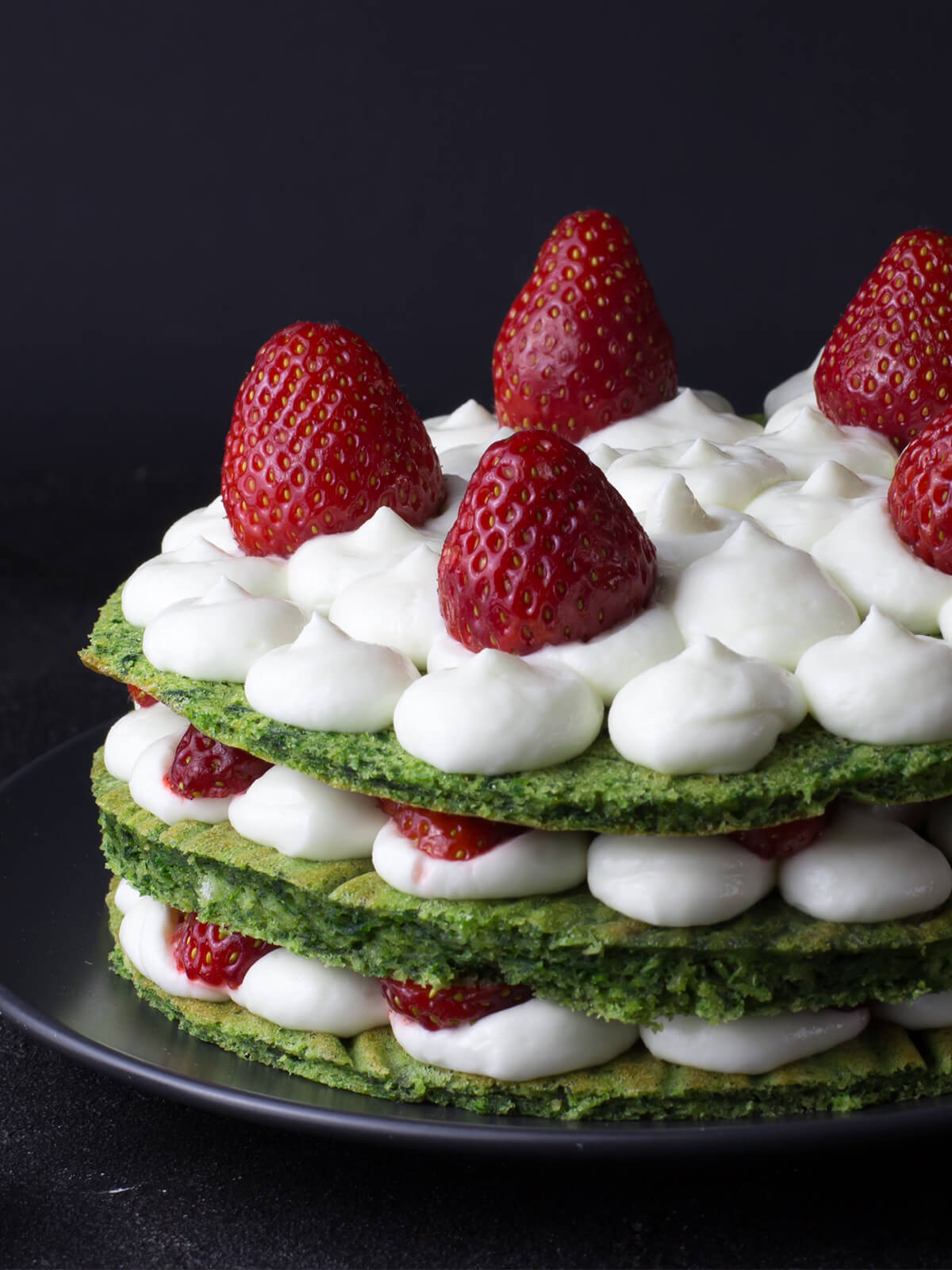 Nahaufnahme der ungeschnittenen grünen Spinat-Torte von der Seite: drei grüne Böden, dazwischen flach gedrückte Sahnetupfen und Erdbeerscheiben