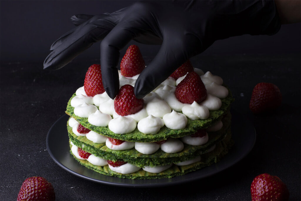 Behandschuhte Hand setzt ganze Erdbeeren auf die Cremetupfen einer grünen Schichttorte