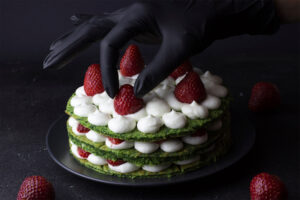 Behandschuhte Hand setzt ganze Erdbeeren auf die Cremetupfen einer grünen Schichttorte
