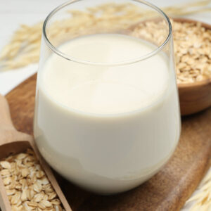 Glas mit cremiger Hafermilch neben einer Schale Haferflocken und Haferrispen