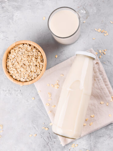 Flasche und Krug mit Hafermilch von oben, daneben eine Holzschale mit Haferflocken