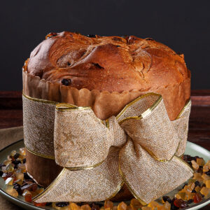 Hoher goldbrauner Panettone in brauner Papierform mit Goldband, rundherum kandierte Fruchtwürfel und Korinthen auf einem hellen Teller