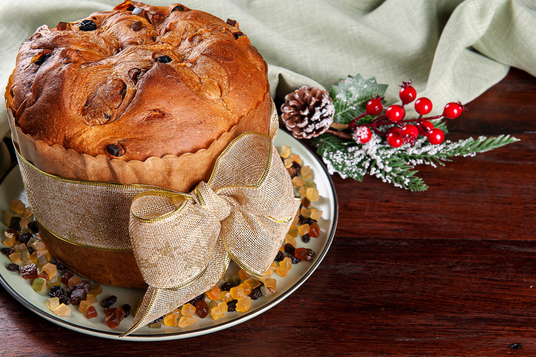 Fertig gebackener Panettone in der Papierform mit Goldband, daneben kandierte Fruchtwürfel, Korinthen und ein Zweig mit roten Beeren