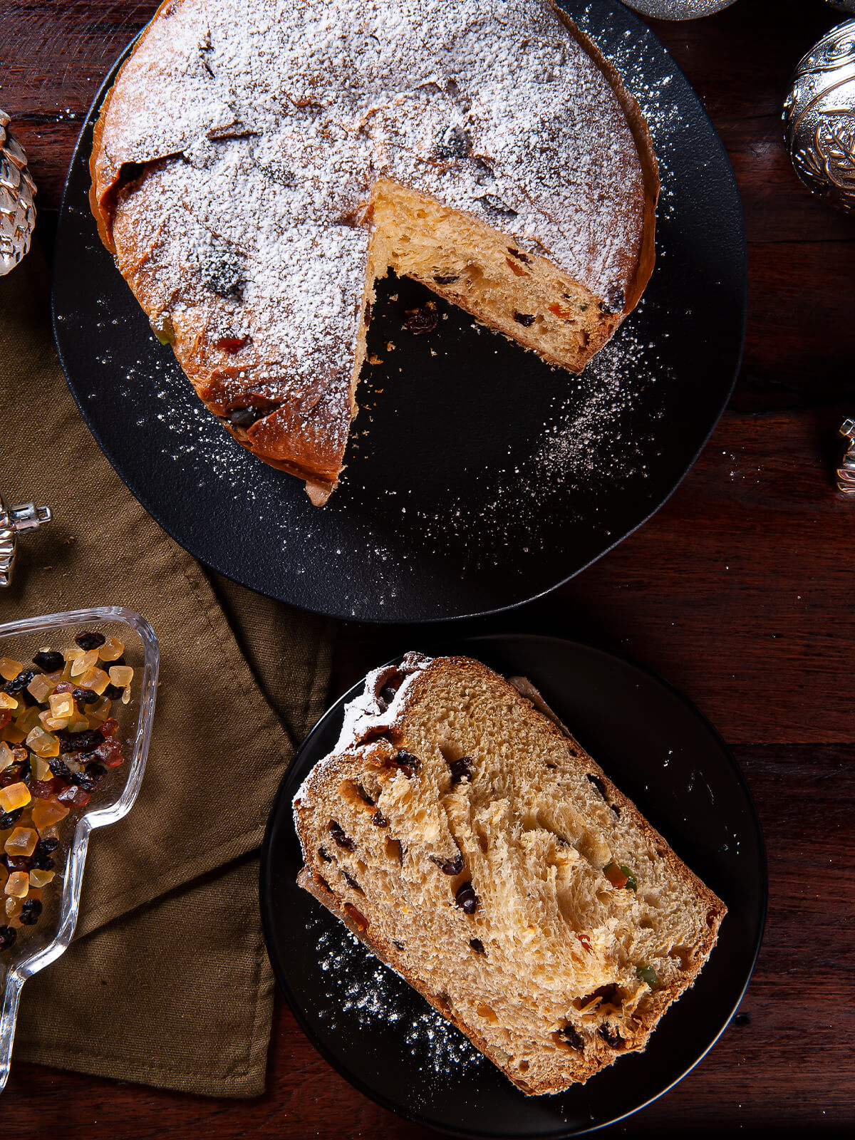 Panettone von oben mit aufgerissenem Kreuzschnitt und Puderzucker, daneben eine dicke Scheibe mit Fruchtstücken auf schwarzem Teller