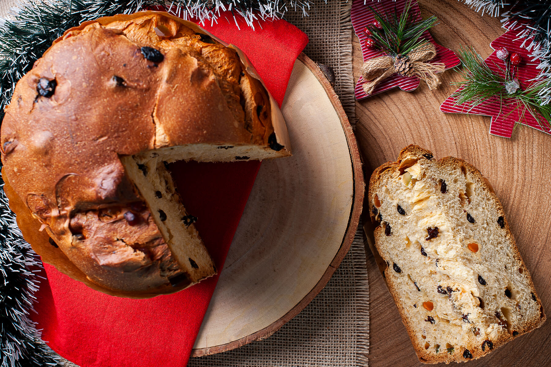 Panettone von oben mit herausgeschnittener Spalte auf rotem Tuch, daneben eine dick mit Butter bestrichene Scheibe auf einem Holzbrett