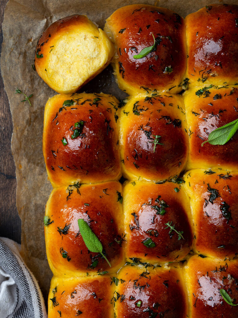 Nahaufnahme eines aufgebrochenen Knoblauchbrötchens mit feiner heller Krume neben glänzenden Brötchen mit Dill-Knoblauch-Butter