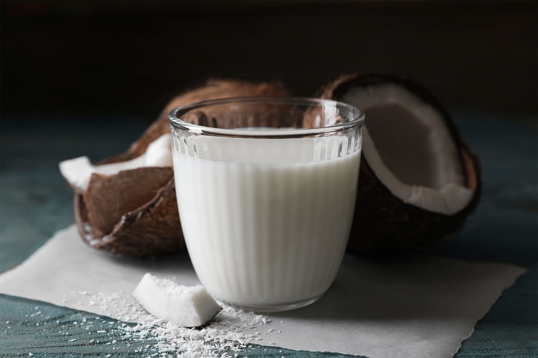 Glas mit dickflüssiger weißer Kokosmilch neben aufgebrochener Kokosnuss und feinen Raspeln
