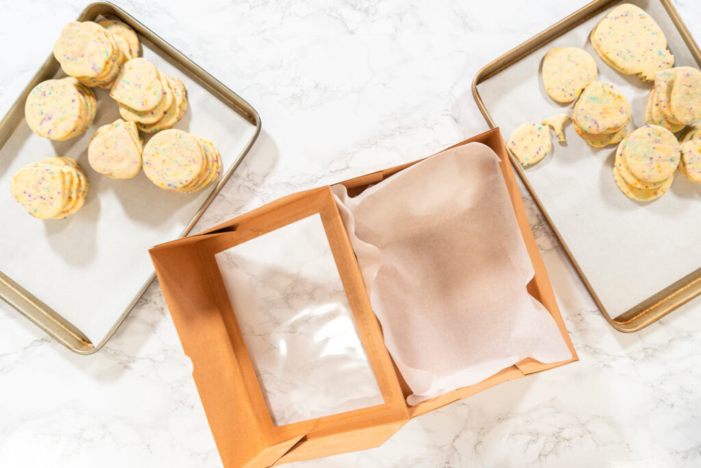 Leere Geschenkschachtel mit Backpapier ausgelegt neben zwei Blechen mit bunten Konfetti-Plätzchen