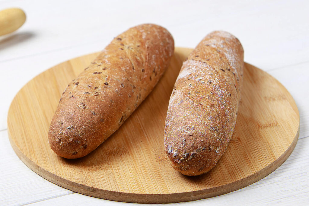 Zwei goldbraune Leinsamen-Baguettes mit Sauerteig nebeneinander auf einem runden Holzbrett