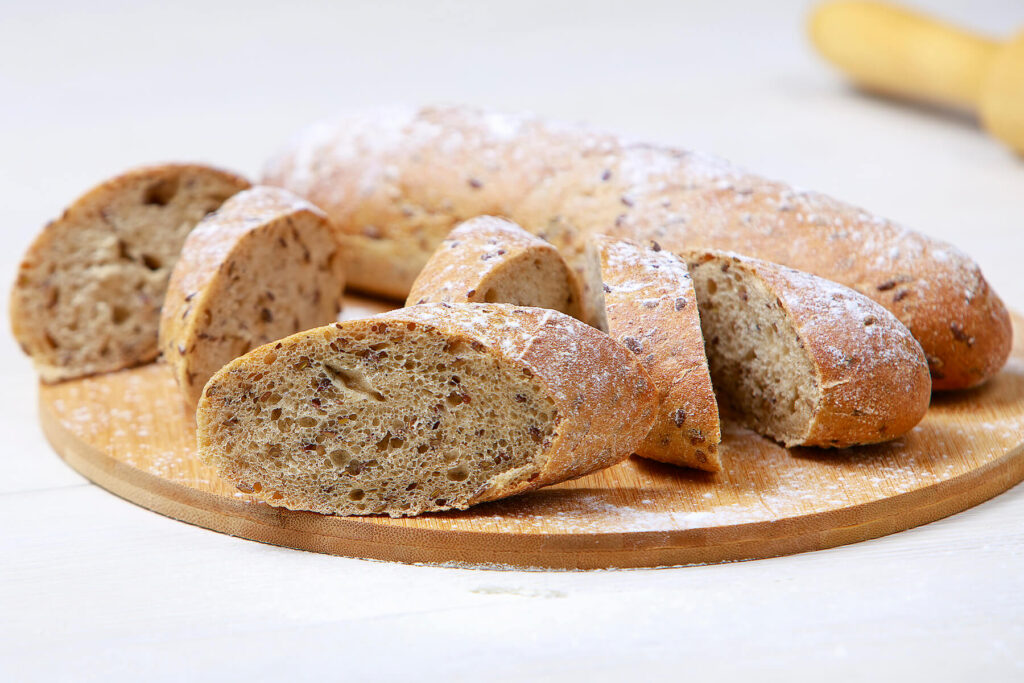 Leinsamen-Baguette mit Sauerteig schräg in dicke Scheiben geschnitten auf einem runden Holzbrett