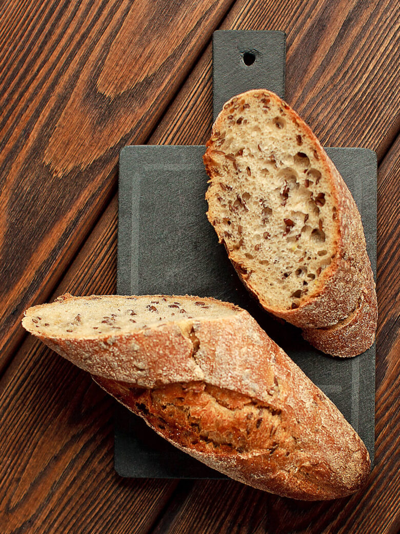 Angeschnittenes Leinsamen-Baguette mit Sauerteig neben einer ganzen Stange auf einer Schieferplatte