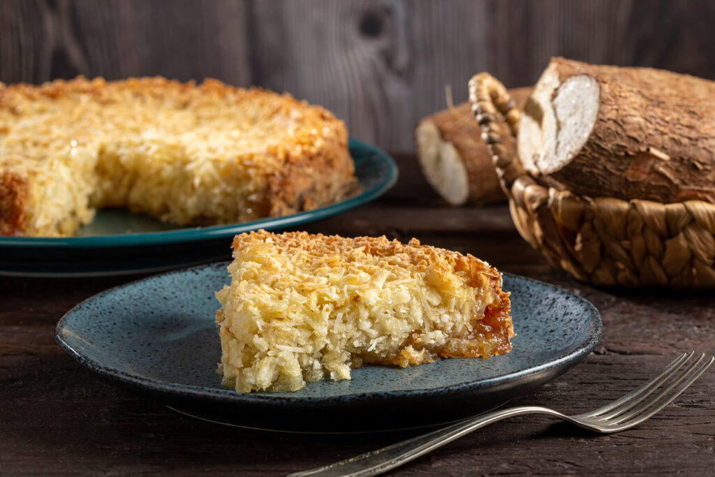 Maniok-Kuchen mit Kokos in der Springform angeschnitten, daneben ein Stück auf blauem Teller