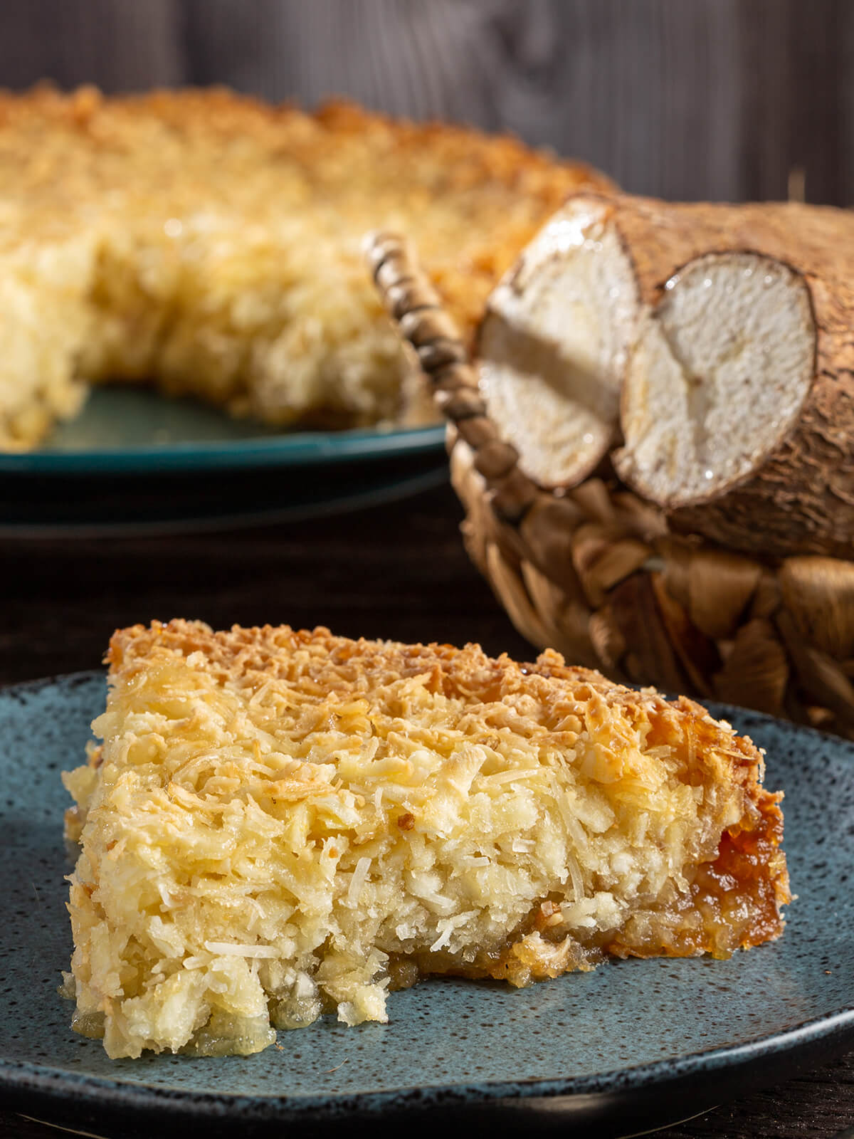 Angeschnittener Maniok-Kuchen mit Kokos, ein Stück auf blauem Teller und frische Maniokwurzel im Korb