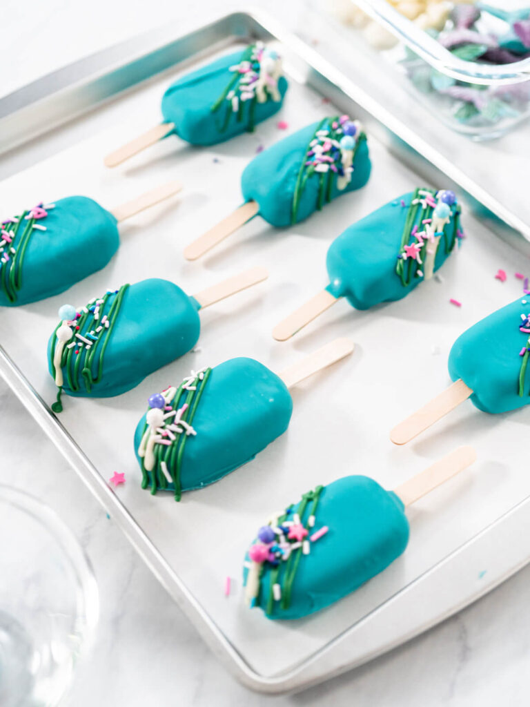Acht getauchte Cakesicles auf dem Blech mit grünen Schokofäden und bunten Streuseln