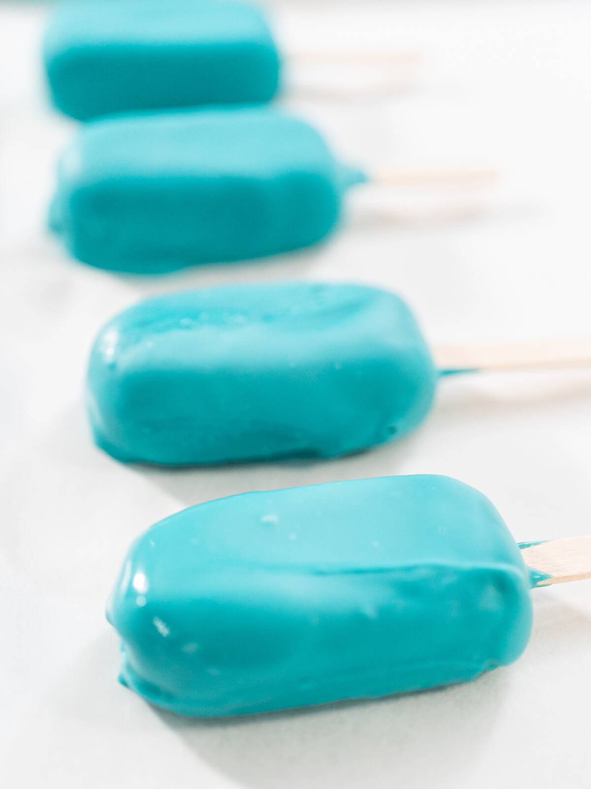 Vier frisch getauchte Cakesicles mit glänzender türkiser Glasur auf Backpapier