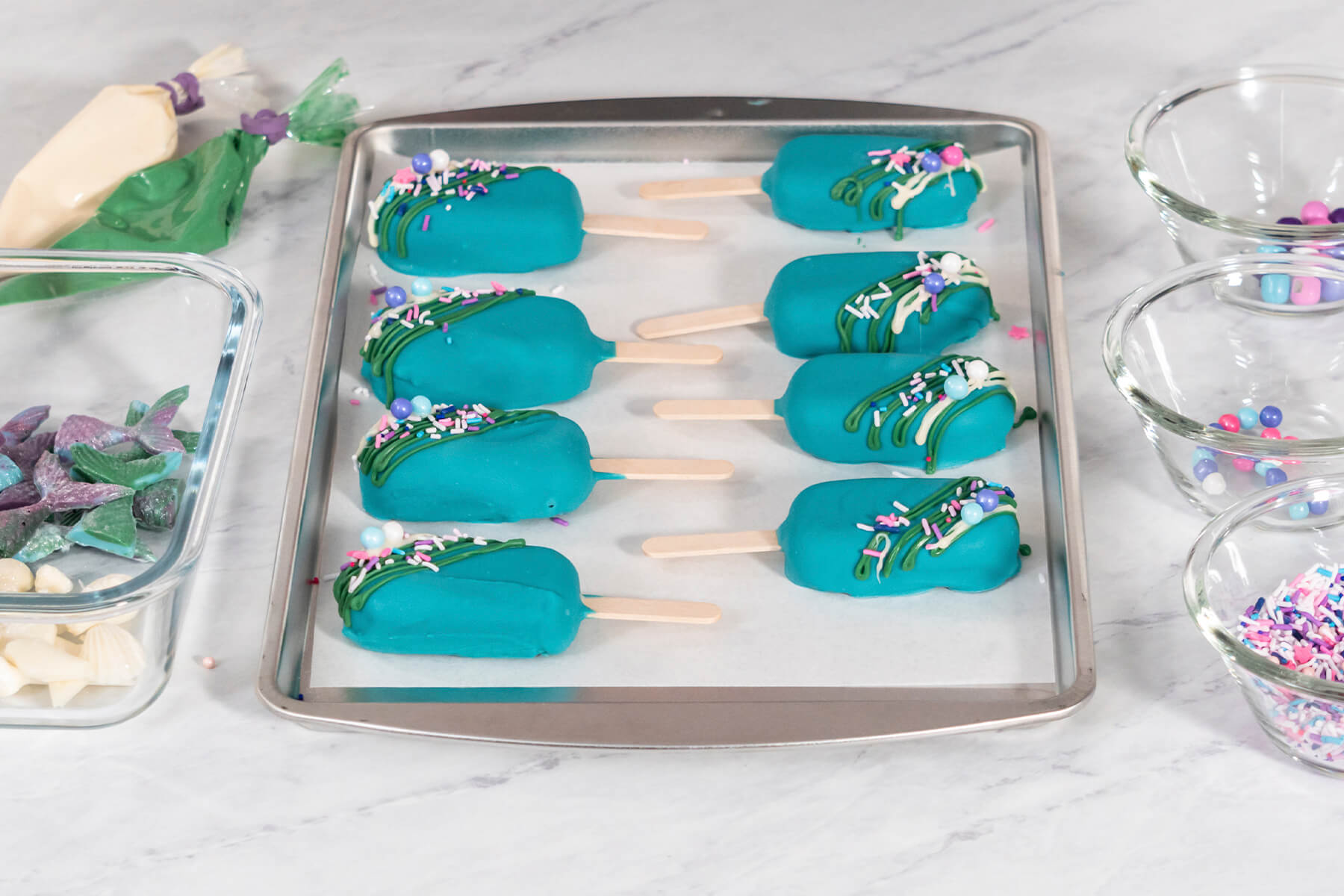 Arbeitsplatz mit dekorierten Cakesicles auf dem Blech, Spritzbeuteln, Schokoflossen und Streuselschalen