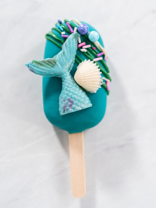 Meerjungfrauen-Cakesicle von oben mit türkiser Flosse, weißer Schokomuschel und grünen Schokofäden