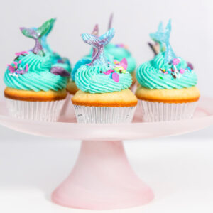Meerjungfrauen-Cupcakes mit türkiser Buttercreme und schimmernden Flossen auf einer rosa Tortenplatte
