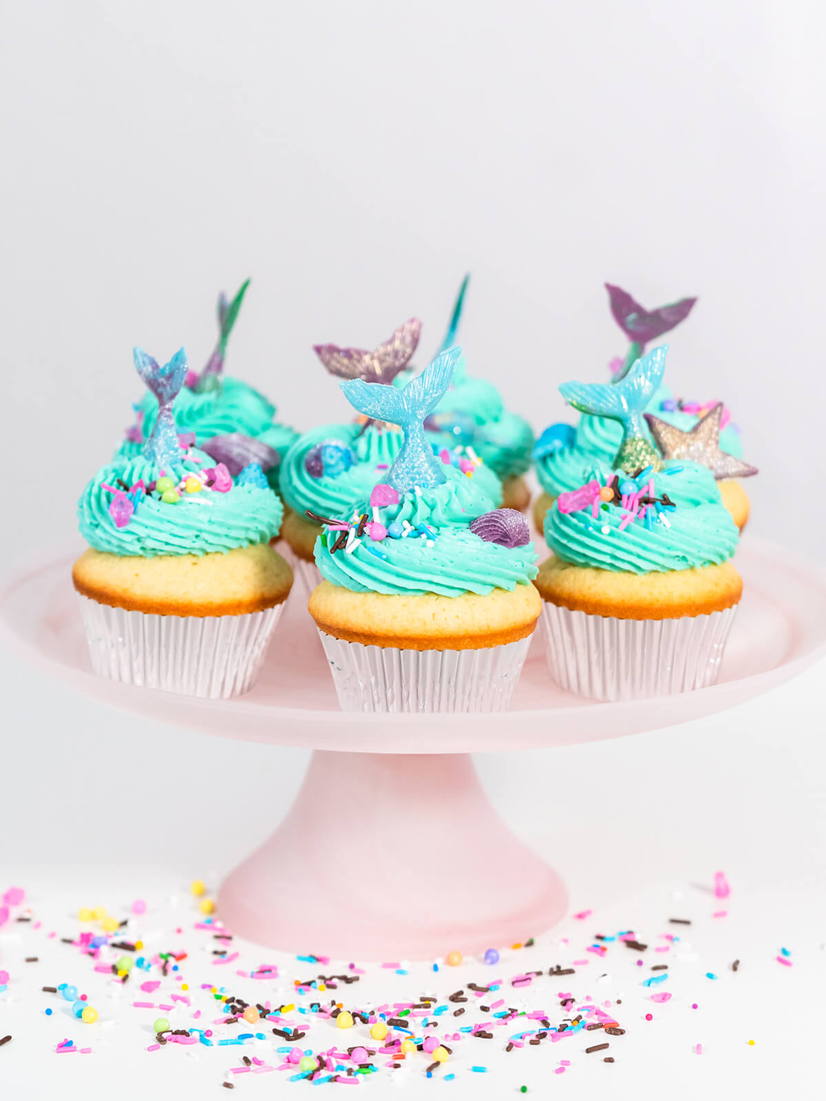 Meerjungfrauen-Cupcakes mit türkiser Buttercreme und schimmernden Flossen auf einer rosa Tortenplatte