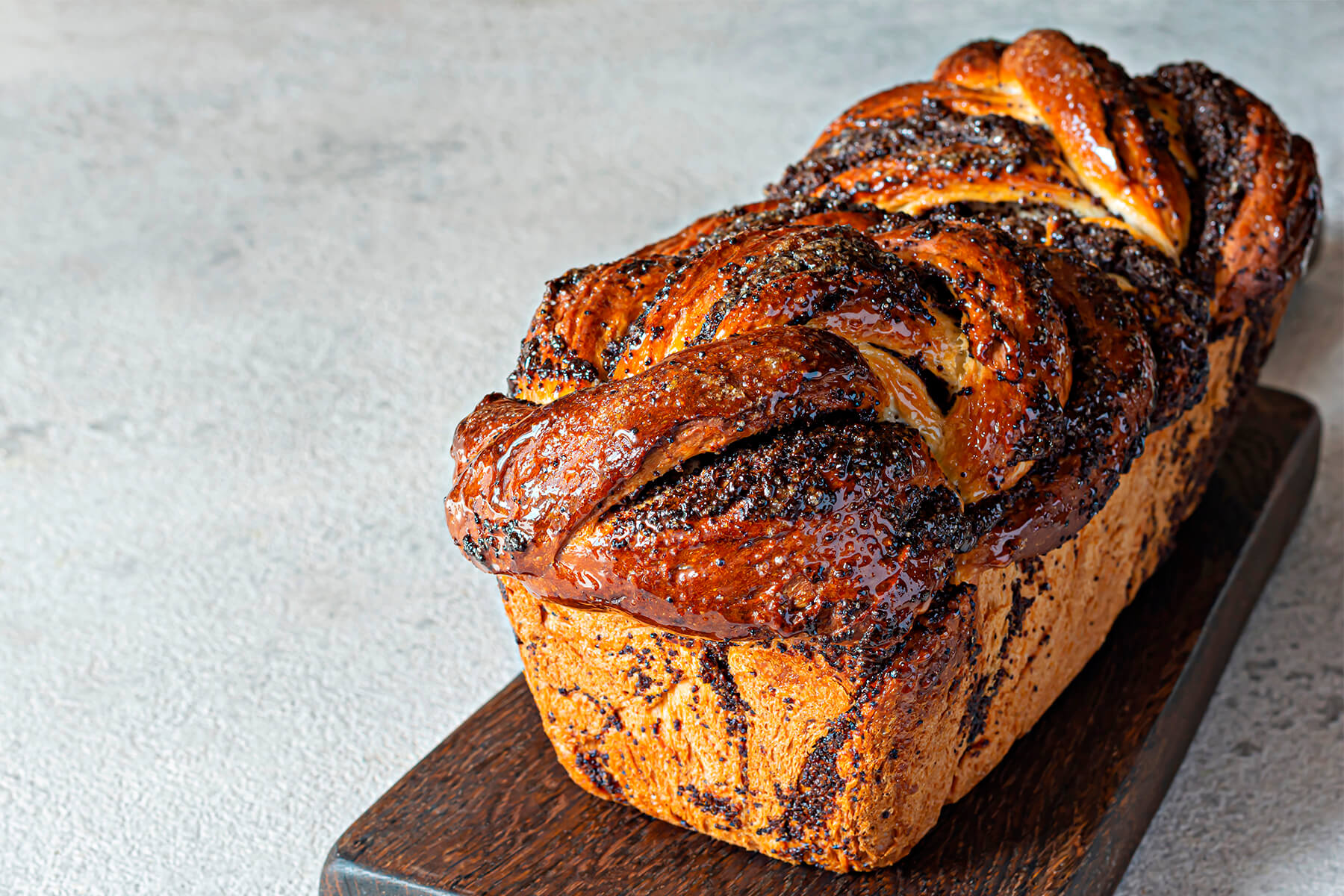 Geflochtene Mohn-Babka mit spiegelnder Zuckersirup-Glasur auf einem dunklen Holzbrett