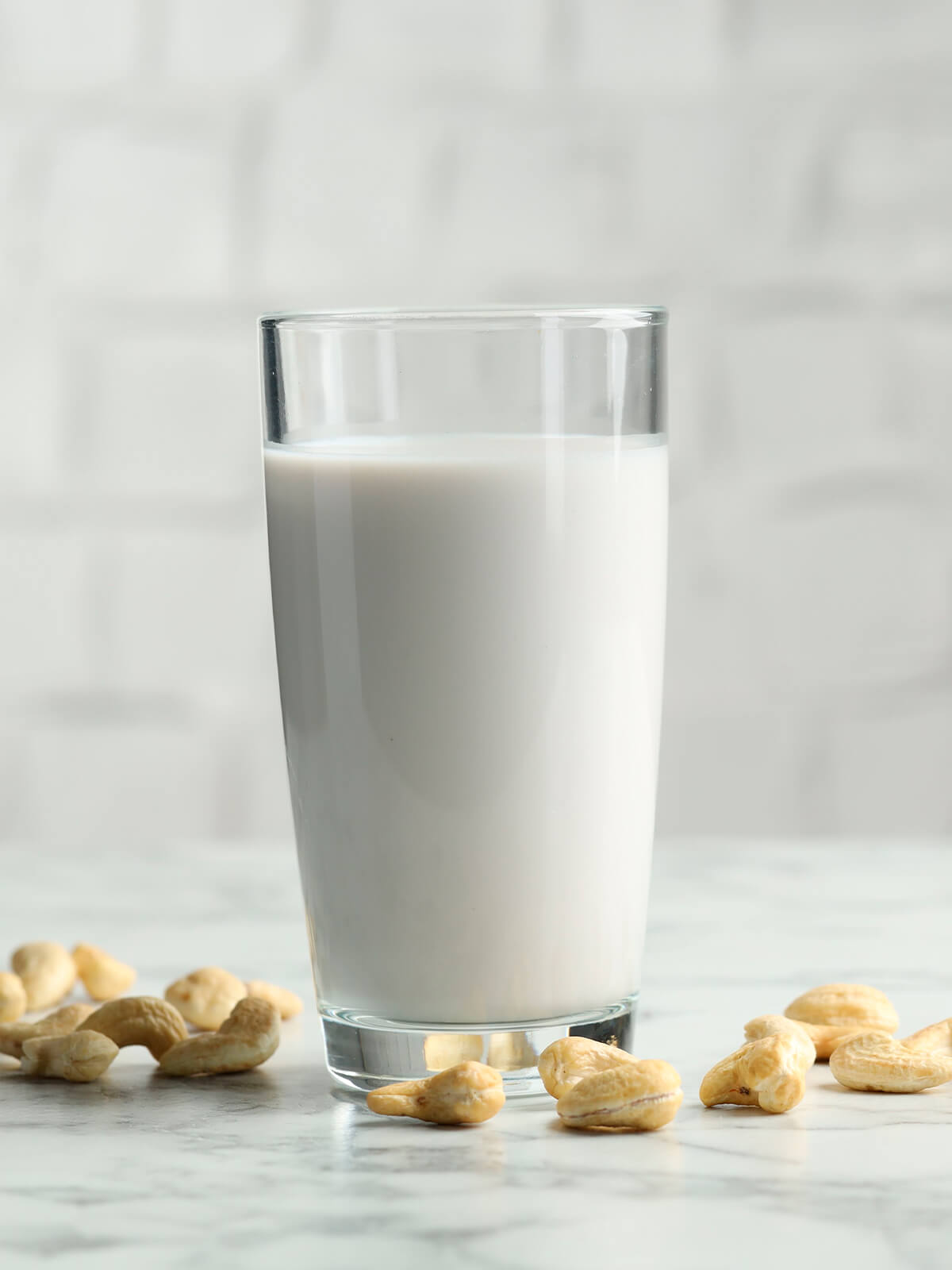 Glas mit frisch abgeseihter Cashewmilch auf hellem Marmor, daneben liegen rohe Cashewkerne