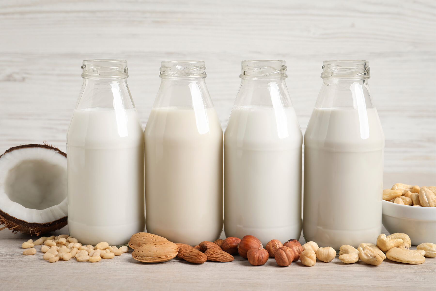 Vier Flaschen mit pflanzlicher Milch aus verschiedenen Nüssen nebeneinander, davor Pinienkerne, Mandeln, Haselnüsse und Cashewkerne