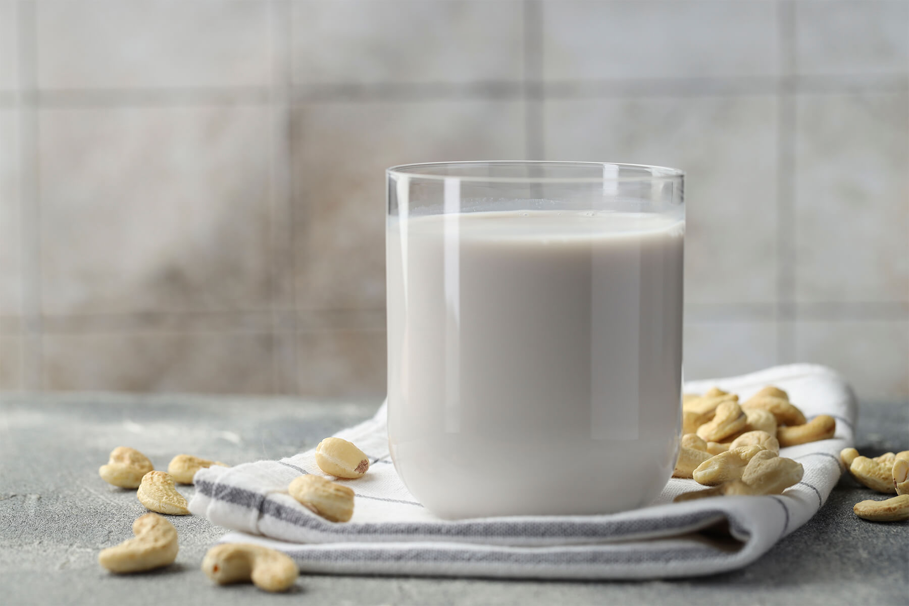 Glas mit cremig-weißer Nussmilch auf einem Geschirrtuch, ringsum verstreute Cashewkerne