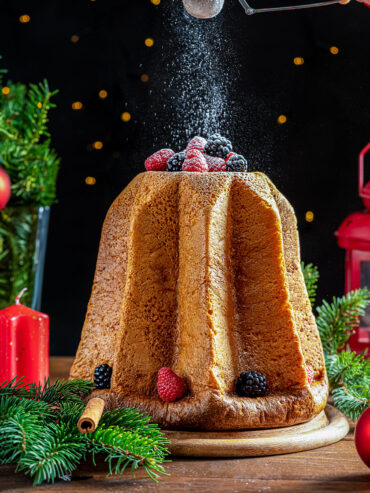 Goldbrauner Pandoro in Sternform auf einem Holzbrett, darüber rieselt Puderzucker aus einem Sieb, obenauf frische Himbeeren und Brombeeren