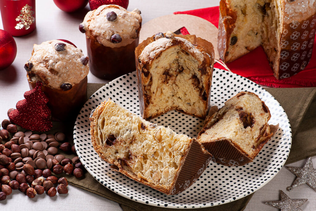 Aufgeschnittene Panettone-Stücke mit Nougat-Spirale auf einem gepunkteten Teller, dahinter ganze Laibe mit Nussglasur