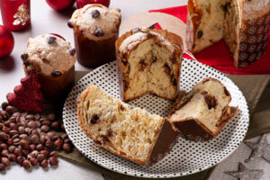 Aufgeschnittene Panettone-Stücke mit Nougat-Spirale auf einem gepunkteten Teller, dahinter ganze Laibe mit Nussglasur