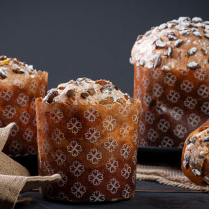 Drei hohe Panettone in gemusterten Papierformen mit heller Pistazienglasur, ganzen Pistazien und Puderzucker vor dunklem Hintergrund