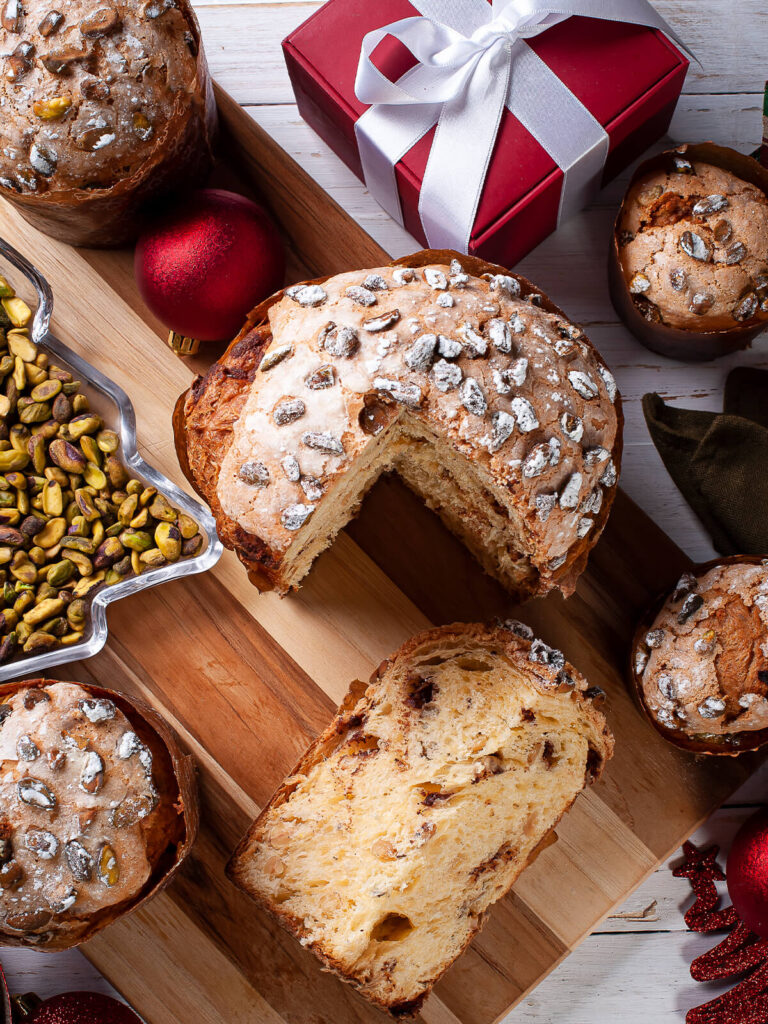 Angeschnittener Pistazien-Panettone auf einem Holzbrett, davor eine dicke Scheibe mit hellen Schokoladennestern