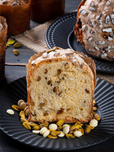 Dicke Scheibe Panettone mit heller Krume, ganzen Pistazien und geschmolzenen Nestern aus weißer Schokolade auf einem schwarzen Teller