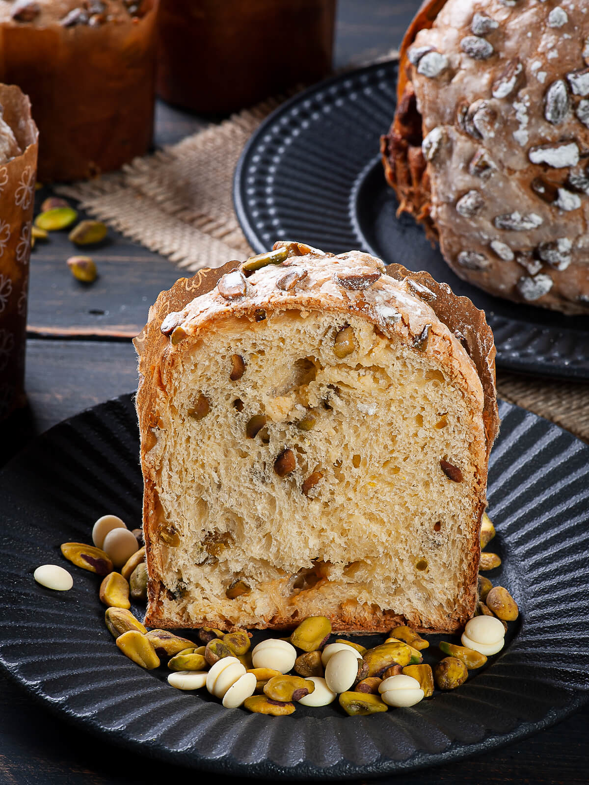 Dicke Scheibe Panettone mit heller Krume, ganzen Pistazien und geschmolzenen Nestern aus weißer Schokolade auf einem schwarzen Teller