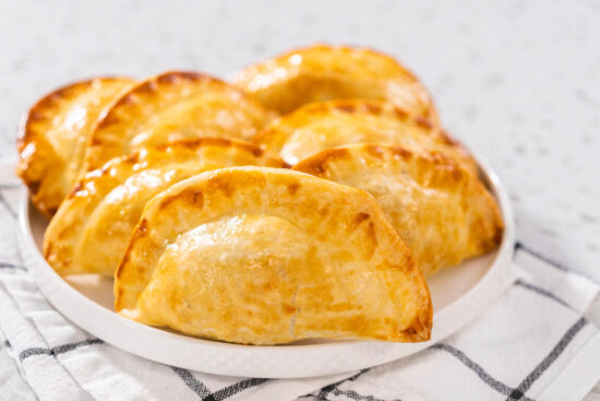 Gebackene Puten-Empanadas mit geriffeltem Rand auf einem weißen Teller, dahinter ein kariertes Geschirrtuch