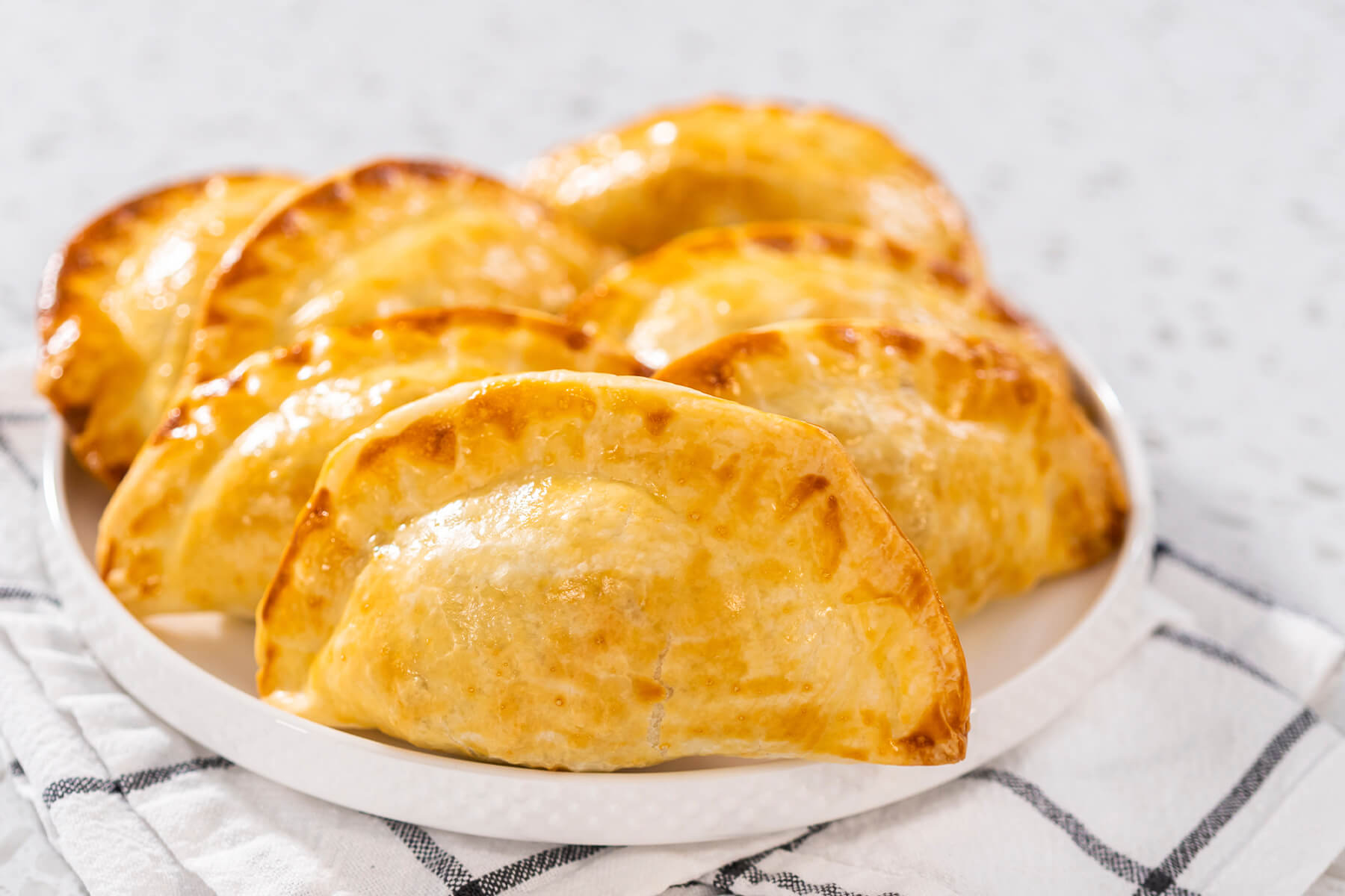 Gebackene Puten-Empanadas mit geriffeltem Rand auf einem weißen Teller, dahinter ein kariertes Geschirrtuch